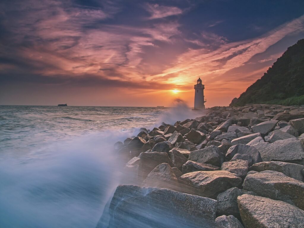 The sun sets beautifully behind a seaside lighthouse, creating a dramatic landscape view.